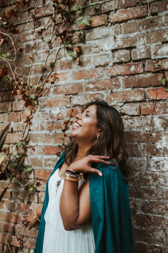 Nisha, wearing a white dress and a green shawl, stands against a brick wall with one hand on her shoulder and smiles warmly.