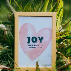 A "Joy is choosing to believe in me" postcard lying in the grass.
