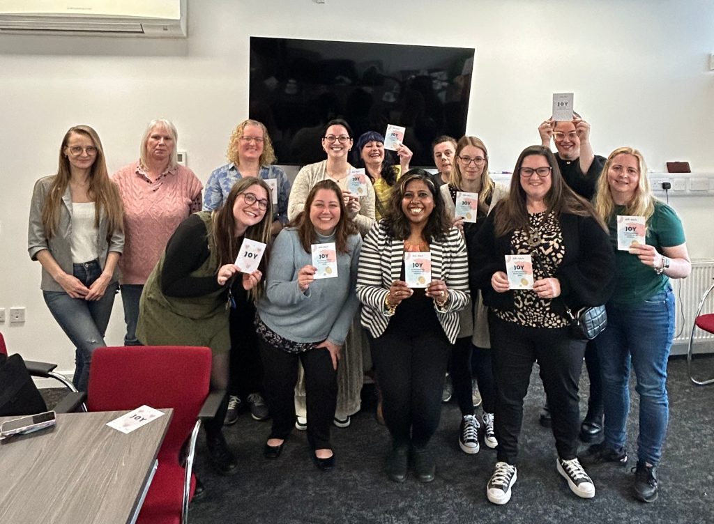 Happy workshop attendees smiling and holding up a Joy postcard while standing with Nisha.